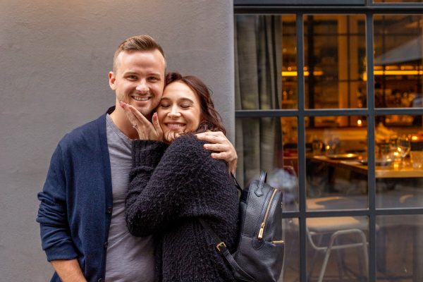 The happy couple sharing a laugh and hugging each other outside a café directly after their engagement.