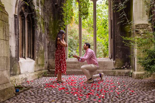 A man down on one knee proposing to his partner on a stone flor at St Dunstan in the East Church Garden