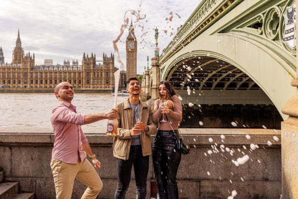 A friend opens a bottle of Champagne after the couple proposal in front of Big Ben at the famous arch