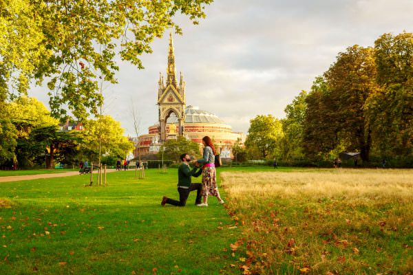 A man down on his knee proposing to his girlfriend in front of Albert Hall in Hyde Park.