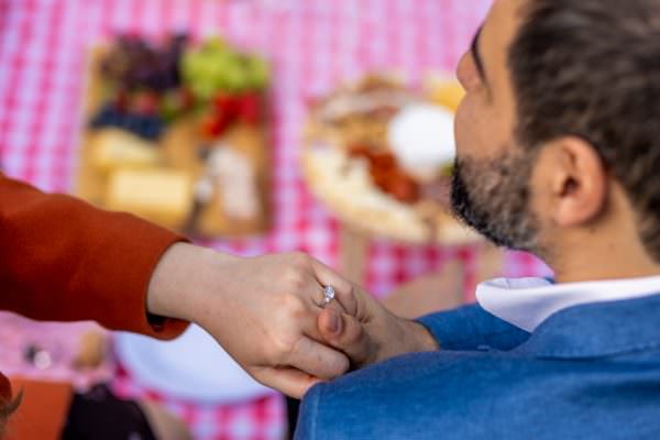A close-up of a sparkling diamond engagement ring on a woman's hand, with a blurred picnic setup in the background at Hyde Park.