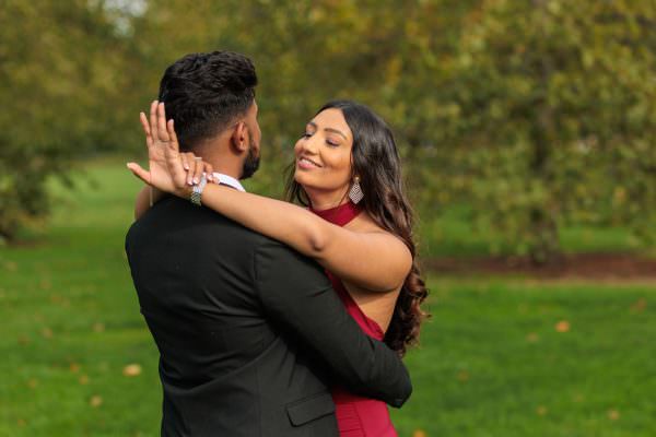 A joyful bride-to-be looking at her new engagement ring as her partner hugs her tightly amidst the greenery of Kensington Gardens, London.