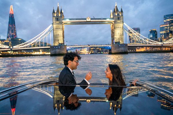 A man standing and proposing to his girlfriend on a boat on the River Thames, holding up an engagement ring with Tower Bridge in the background.